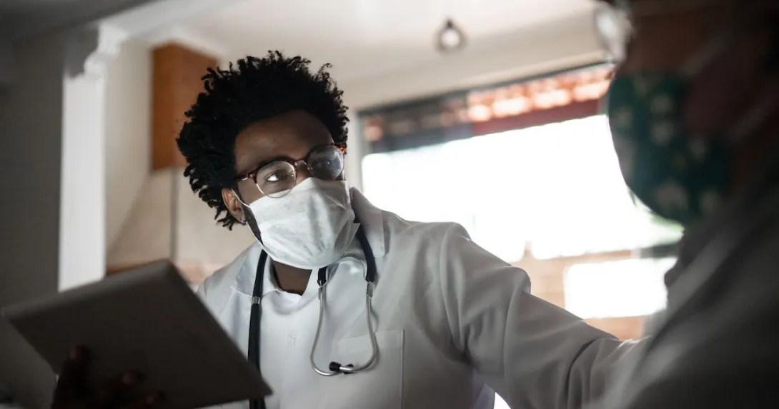 Healthcare provider wearing a mask, a lab coat and a stethoscope around their neck talking to a patient in their home Healthcare provider wearing a mask, a lab coat and a stethoscope around their neck talking to a patient in their home