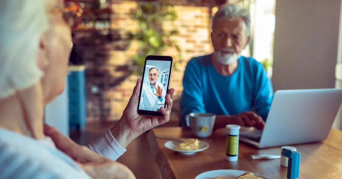 Two people sitting at a table with a computer on it and one of them is holding a phone Two people sitting at a table with a computer on it and one of them is holding a phone