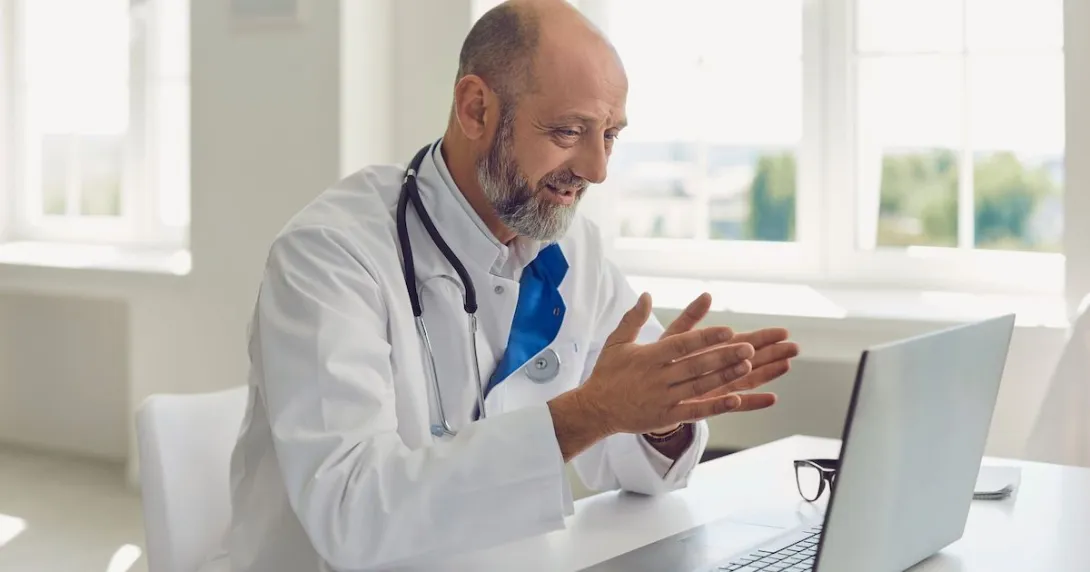 Healthcare provider sitting at a desk, wearing a lab coat and stethoscope, while looking at a computer Healthcare provider sitting at a desk, wearing a lab coat and stethoscope, while looking at a computer