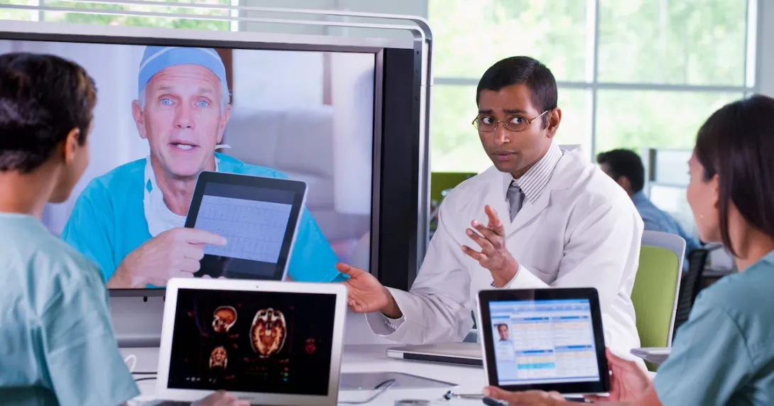 Healthcare team sitting around a table with laptops on it while talking to another healthcare provider on a TV screen