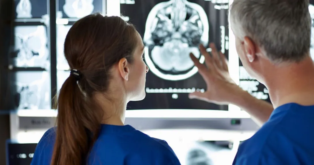 Two healthcare providers looking at a diagnostic image of a brain on a wall Two healthcare providers looking at a diagnostic image of a brain on a wall