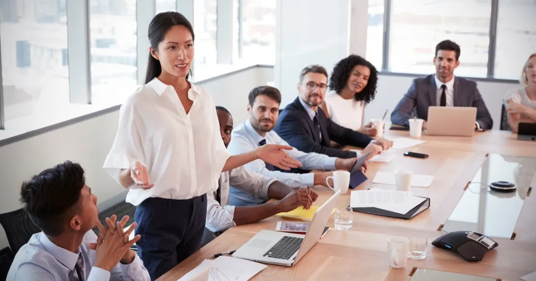 Numerous people in a board room sitting around a table with one person standing up Numerous people in a board room sitting around a table with one person standing up