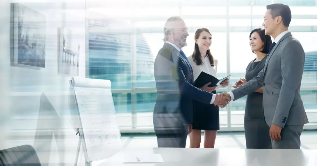 Four people standing in a room with two of them shaking hands Four people standing in a room with two of them shaking hands