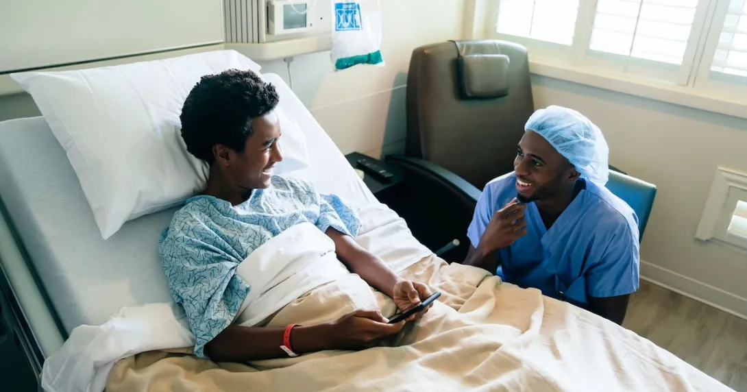 Healthcare provider sitting next to a hospital bed with a teenager in it looking at a phone Healthcare provider sitting next to a hospital bed with a teenager in it looking at a phone