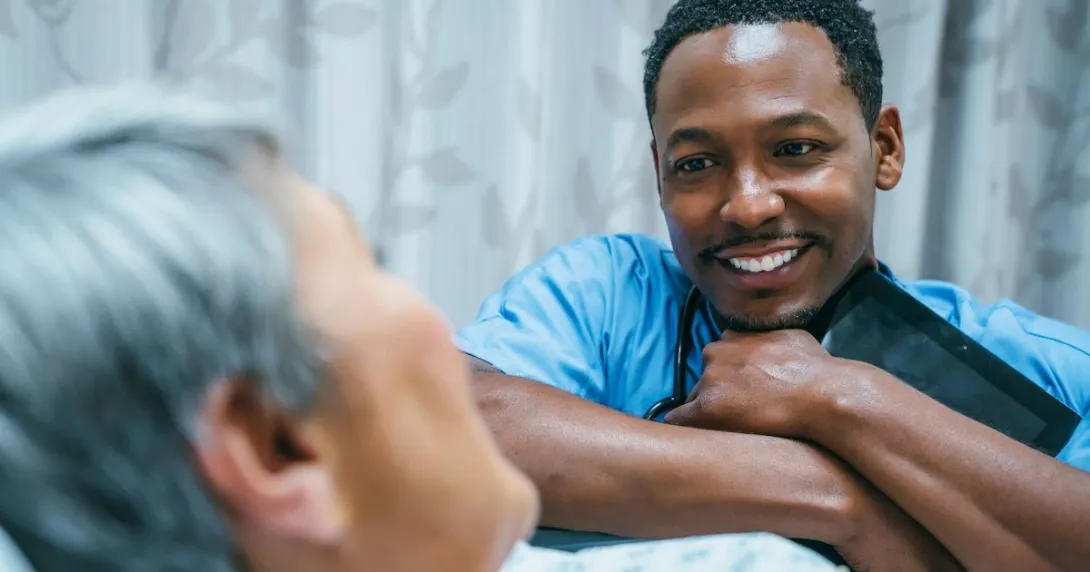 Nurse leaning against a patient's bed while the patient looks at them Nurse leaning against a patient's bed while the patient looks at them